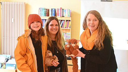 Zwei Frauen und ein Mädchen stehen gemeinsam in einem größeren Raum. Ein Bücherregal ist im Hintergrund zu sehen. Das Mädchen und eine der Frauen halten jeweils ein Stofftier in den Händen.