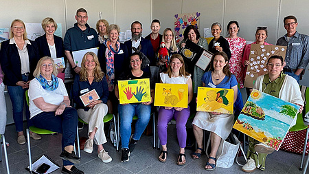 Ein größere Gruppe Frauen und Männer steht oder sitzt in einem Raum. Einige halten Bilder in Händen.
