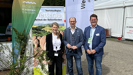 Eine Frau und zwei Männer stehen vor mehreren Werbebannern.