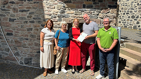 Drei Frauen und zwei Männer stehen in Freien vor einer Steinmauer und einem Treppenaufgang. Eine der Frauen überreicht einem der Männer ein Dokument.