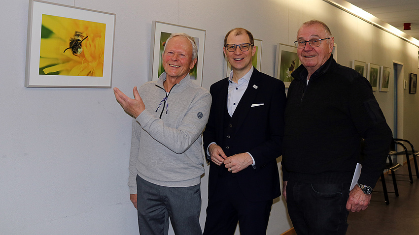 Drei Männer stehen nebeneinander in einem Flur. Der Mann ganz links deutet auf eine Fotografie, die an der Wand hängt.