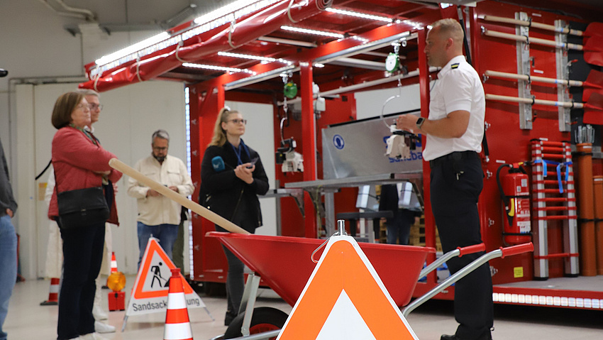Ein Feuerwehrmann erläutert mehreren Personen die Funktionsweise von Feuerwehrausrüstung. Diese ist im Hintergrund zu sehen.