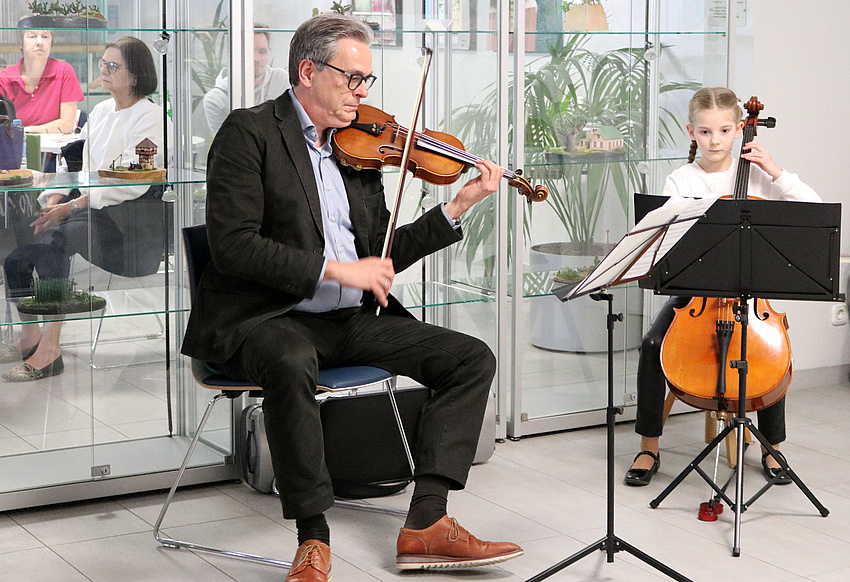 Ein Mann spielt Violine. Rechts neben ihm spielt ein Mädchen Cello. Beide haben Notenständer vor sich.