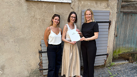 Drei Frauen stehen vor einer Gebäudewand. Eine der Frauen übergibt ein Dokument an eine andere.