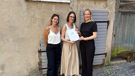 Drei Frauen stehen vor einer Gebäudewand. Eine der Frauen übergibt ein Dokument an eine andere.