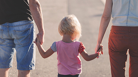 Zwei Erwachsene halten ein kleines Kind an den Händen, die drei Personen sind von hinten zu sehen.