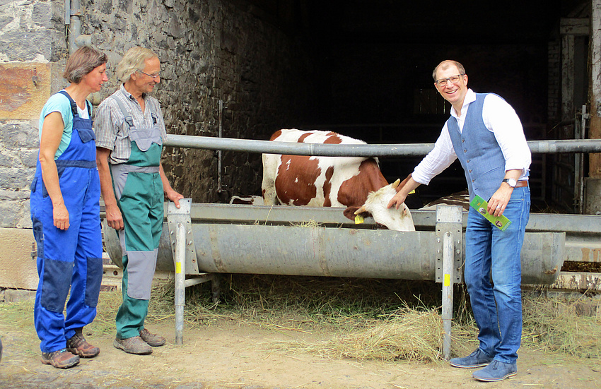 Drei Personen auf einem Bauernhof. Hinter einer Absperrung eine Kuh, die aus einem metallenen Futtertrog frisst