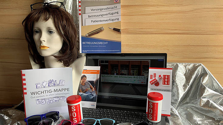 Auf einem Tisch liegen Brillen, Handys, ein Kopf mit Perücke steht links.