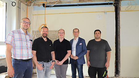 Fünf Männer stehen in einem Raum, in dem Bauarbeiten stattfinden. Kabel hängen von der Decke und in den Wänden sind offene Kabelschächte zu sehen.