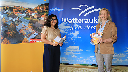 Zwei Frauen vor einem Aufsteller mit der Aufschrift "Wetteraukreis". Sie zeigen Flyer für die Energieberatung.