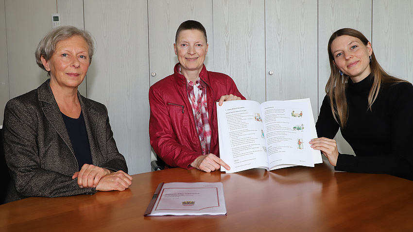 Wera-Marianne Arndt, Marion Götz und Anaïs Hoffmann präsentieren an einem Tisch den Aktionsplan Inklusion.