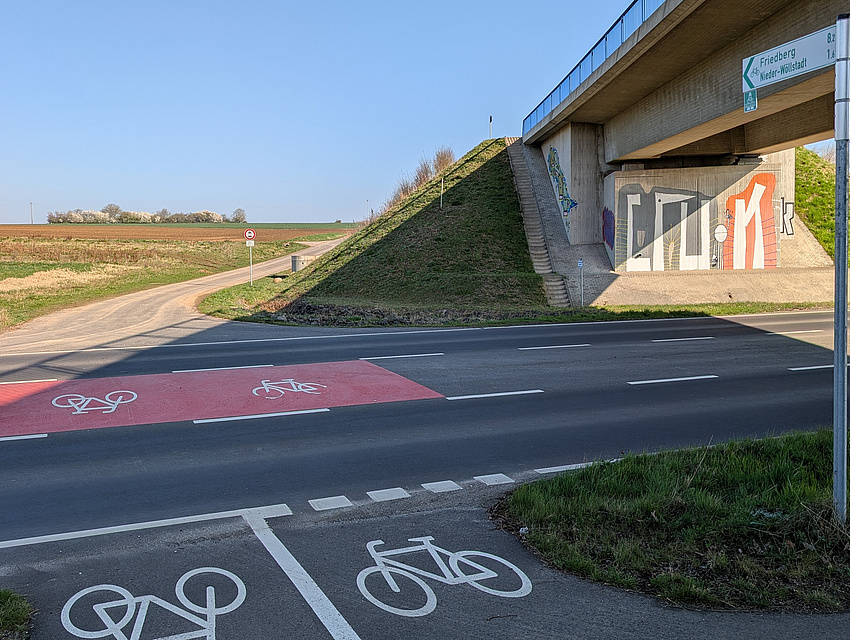 Blick auf eine rote Markierung mit Fahrradsymbolen auf der Fahrbahn, die Radfahrern helfen soll, eine Straße sicher zu überqueren. Am rechten Bildrand ist eine Brücke zu sehen.