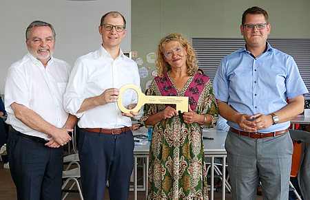 Drei Männer und eine Frau stehen nebeneinander. Die Frau und der Mann in der Mitte halten gemeinsam einen großen symbolischen Schlüssel
