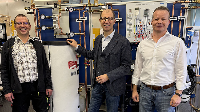 Drei Männer stehen in einem Werkstattraum vor mehreren technischen und elektronischen Apparaturen.