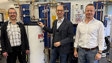 Drei Männer stehen in einem Werkstattraum vor mehreren technischen und elektronischen Apparaturen.