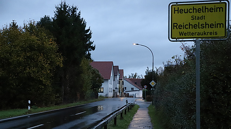 Eine regenasse Straße führt an einem Ortsschild vorbei in ein Dorf. Der Himmel ist bedeckt und die Straßenlaternen leuchten noch.