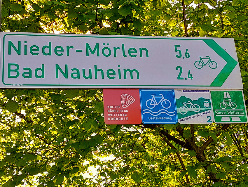 Radneubeschilderung - Blick auf ein richtungsweisendes Schild mit Kilometerangaben und vier Logo-Einschubplaketten, die auf diverse Themen-Radwege hinweisen.