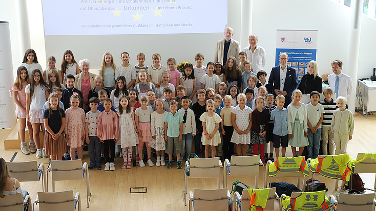 Zahlreiche jüngere Schülerinnen und Schüler stehen gemeinsam mit einigen Erwachsenen in einem großen Saal. Im Hintergrund sind ein Plakat und der Start einer PowerPoint-Präsentation auf einer Leinwand zu sehen.