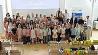 Zahlreiche jüngere Schülerinnen und Schüler stehen gemeinsam mit einigen Erwachsenen in einem großen Saal. Im Hintergrund sind ein Plakat und der Start einer PowerPoint-Präsentation auf einer Leinwand zu sehen.