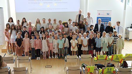 Zahlreiche jüngere Schülerinnen und Schüler stehen gemeinsam mit einigen Erwachsenen in einem großen Saal. Im Hintergrund sind ein Plakat und der Start einer PowerPoint-Präsentation auf einer Leinwand zu sehen.
