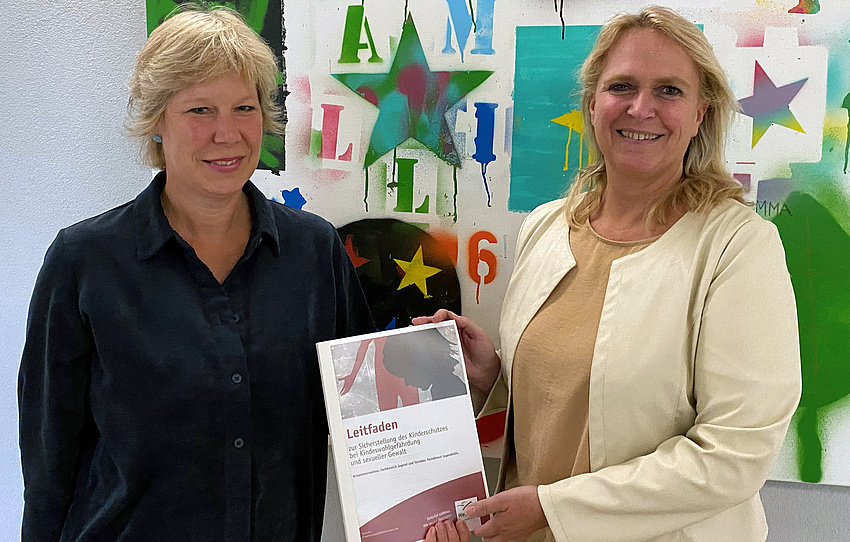 Zwei Frauen stehen nebeneinander. Sie halten gemeinsam ein Schriftstück in der Hand