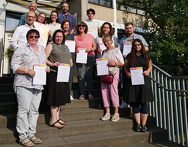 Mehrere Personen stehen auf einer Treppe. Alle haben ein Schriftstück in der Hand.