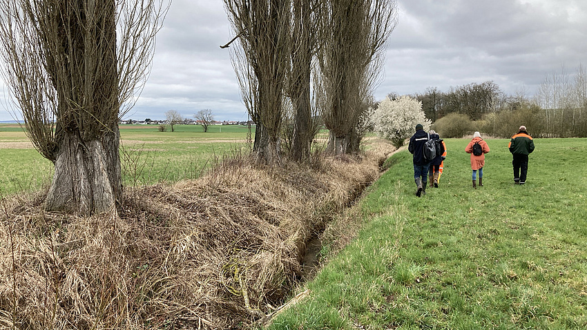 Vier Personen laufen rechts an einem Bachlauf entlang vom Betrachter weg. Auf der linken Seite des Bachs sind mehrere Bäume zu sehen.
