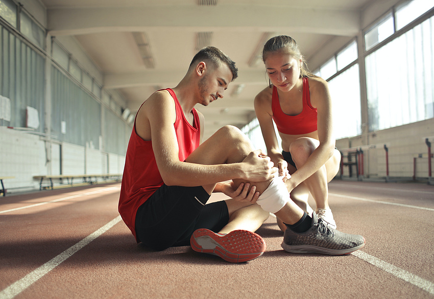In einer Sporthalle: Eine junge Frau verbindet das Bein eines jungen Mannes.
