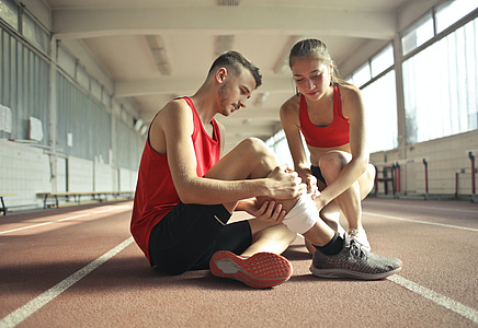 In einer Sporthalle: Eine junge Frau verbindet das Bein eines jungen Mannes.