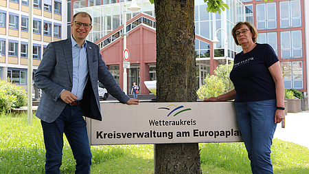 Ein Mann und eine Frau stehen vor einem Baum, um den herum ein Schild mit dem Schriftzug Wetteraukreis angebracht ist.