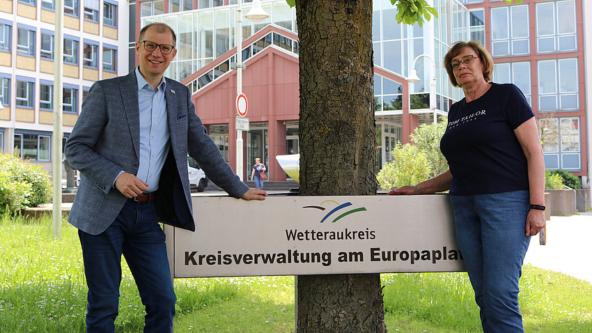 Ein Mann und eine Frau stehen vor einem Baum, um den herum ein Schild mit dem Schriftzug Wetteraukreis angebracht ist.