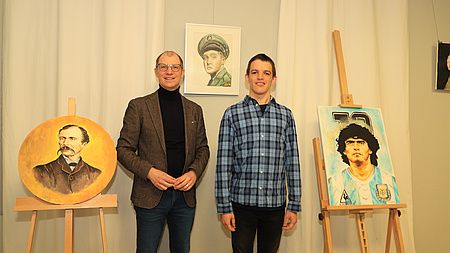 Zwei Männer stehen in einem großen Raum vor einer Wand. Rechts und links von ihnen stehen Gemälde auf Staffeleien, ein weiteres hängt an der Wand.