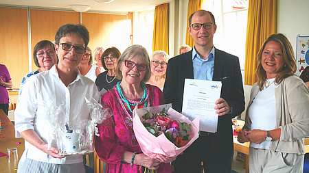 Vier Personen stehen nebeneinander. Die Person in er MItte wird geerht und hält einen Blumenstrauß in der Hand und erhält eine Urkunde.