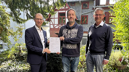 Drei Männer stehen vor einem Fachwerkhaus. Sie halten ein Dokument in den Händen.
