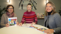 Drei Frauen sitzen in einem Raum gemeinsam an einem Tisch. Vor zweien von ihnen liegt eine Broschüre auf dem Tisch, die dritte hält die gleiche Broschüre in Händen.