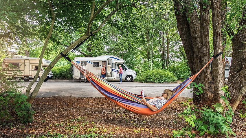 Frau in einer Hängematte im Wald, dahinter parken Wohnmobile