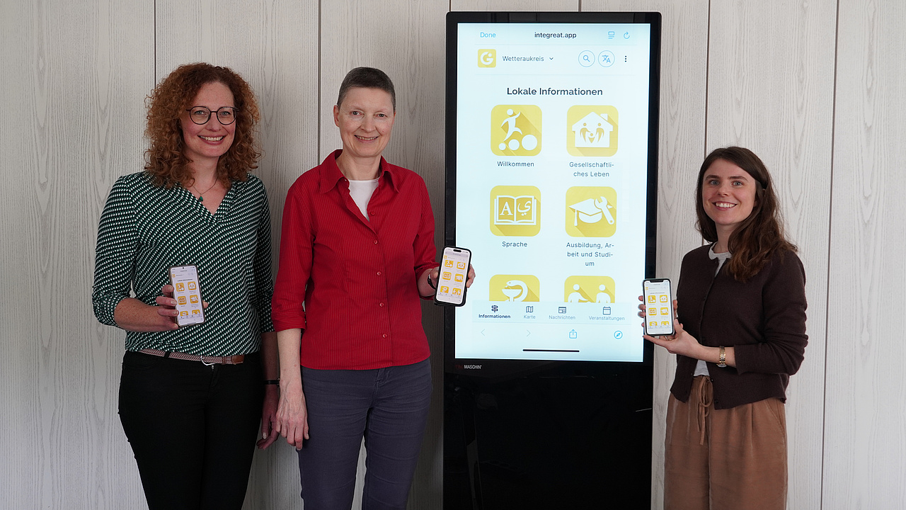 Drei Frauen stehen halten ihr Handy hoch mit der vorgestellen App. Daneben ein Plakat der App.