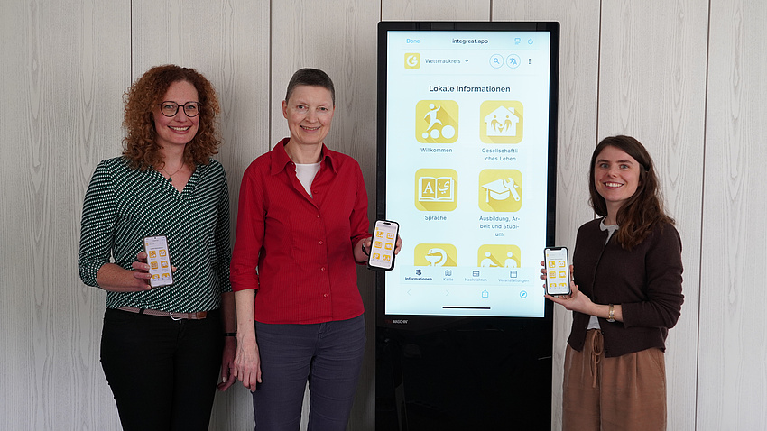 Drei Frauen stehen in einem Raum um einen digitalen Info-Bildschirm und halten jeweils ein Smartphone in die Kamera.