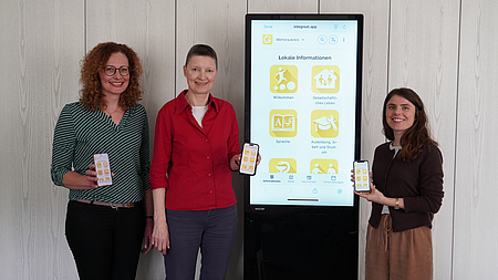Drei Frauen stehen in einem Raum um einen digitalen Info-Bildschirm und halten jeweils ein Smartphone in die Kamera.