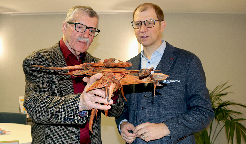 Zwei Männer. Einer hält eine bearbeitete Baumwurzel in der Hand und erklärt dem anderen Mann etwas. Beide schauen die Wurzel an.