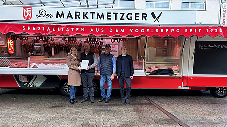 Eine Frau und drei Männer stehen auf einem Platz im Freien vor einem Verkaufswagen mit der Aufschrift einer Metzgerei. Die Frau übergibt einem der Männer ein Dokument.