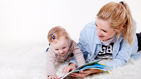 Frau liegt mit ihrem Kleinkind auf einem Teppich und schaut ein Buch an