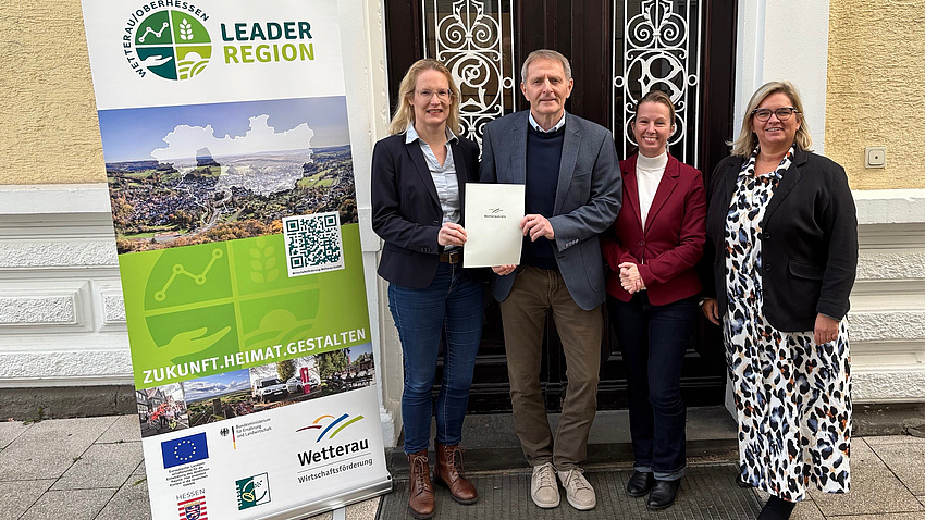 Drei Frauen und ein Mann stehen vor der Tür eines Gebäudes. Eine der Frau und der Mann halten gemeinsam ein Dokument in Händen.
