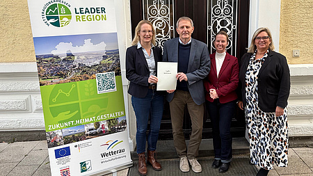 Drei Frauen und ein Mann stehen vor der Tür eines Gebäudes. Eine der Frau und der Mann halten gemeinsam ein Dokument in Händen.