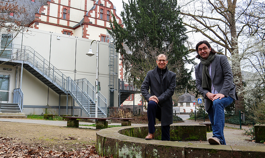 Zwei Männer an einem kleiinen halbrunden Steinmäuerchen. Beide haben jeweils einen Fuß auf das Mäuerchen gestellt. Im Hintergrund ein Schulgebäude.