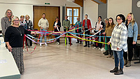 Eine Gruppe von Leuten hat sich im Kreis aufgestellt. Sie halten bunte Bänder in den Händen, die in der Mitte einen Knoten bilden.