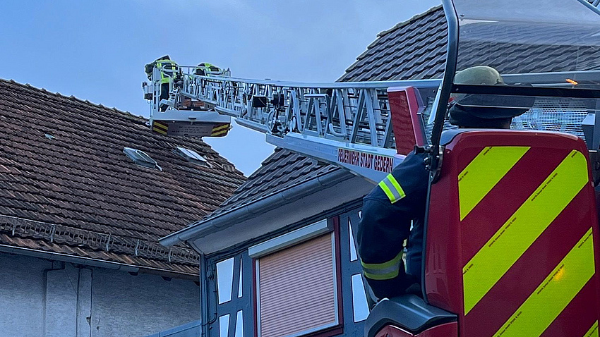 Zu sehen ist eine Feuerwehr-Drehleiter. Feuerwehrleute reparieren ein beschädigtes Dach.