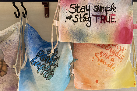 Mehrere bemalte und Besprühte Stoffrucksäcke hängen an einer Garderobe