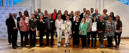 Eine Gruppe von Menschen hat sich um eine Bühne gruppiert. Einige halten ein Zertifikat in der Hand.