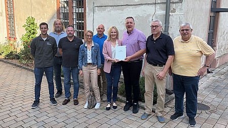 Eine Personengruppe, zwei Frauen und sieben Männer, steht vor einer sanierungsbedürftigen Gebäudefassade. Eine der Frauen und einer der Männer halten gemeinsam ein Dokument in Händen.